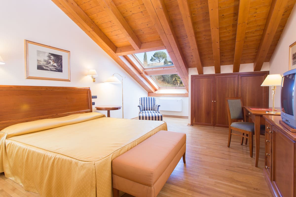 A bedroom with a large bed, a wooden ceiling, a skylight, a desk with a chair, and a television.