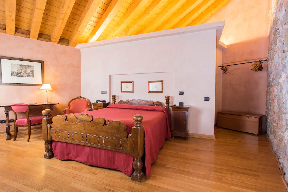 A bedroom with a wooden ceiling, a bed with a red bedspread, a desk with a chair, and a nightstand.