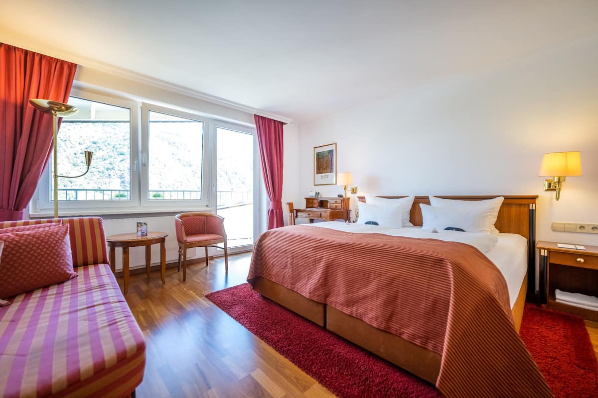 Ein Hotelzimmer mit einem großen Bett, einem Schreibtisch, einem Stuhl und Blick ins Freie durch das Fenster.