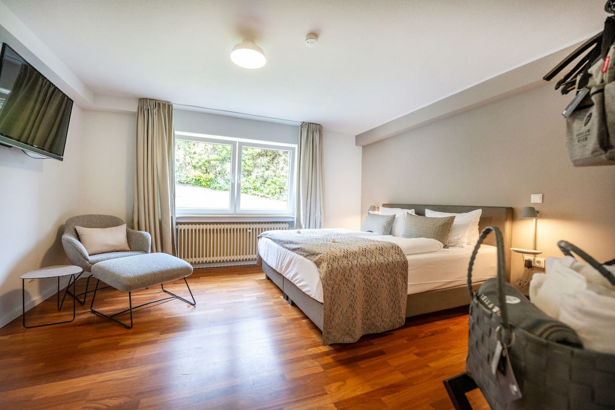 Ein Hotelzimmer mit Bett, Sessel, Fenster, Heizkörper und Wandfernseher.