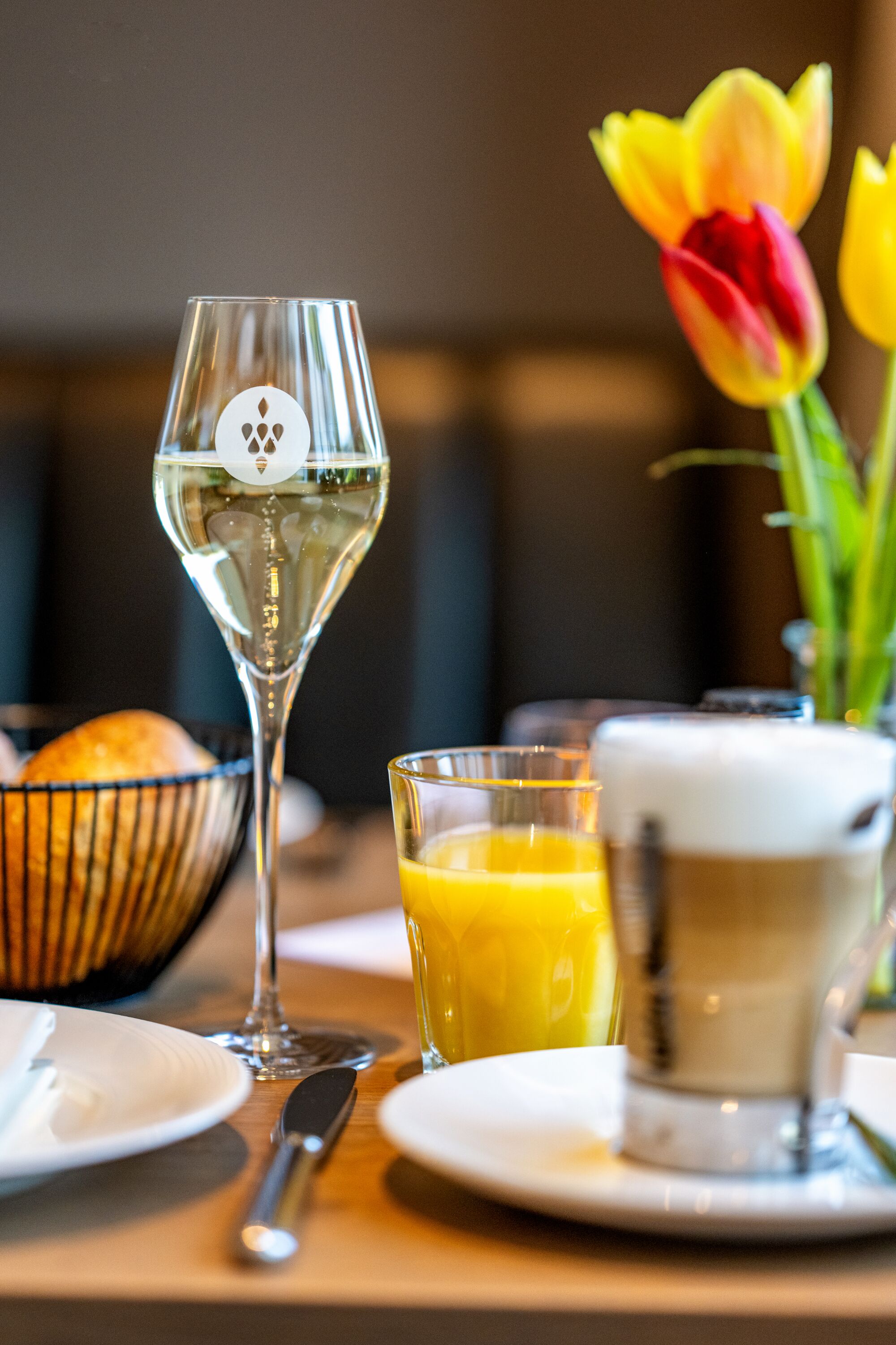 A dining table with a wine glass, a glass of orange juice, a cup of coffee, a bowl of bread, and a tulip.