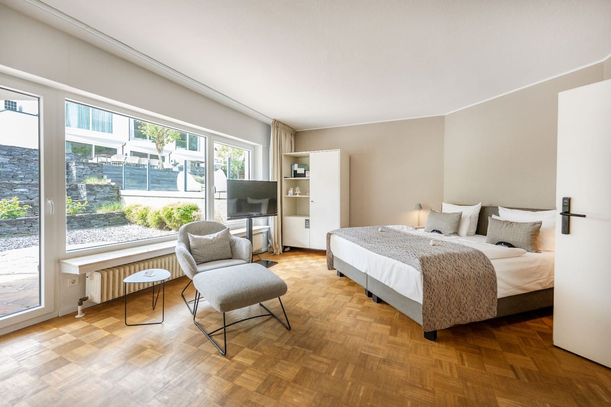 Ein modernes Hotelzimmer mit einem großen Bett, einem Fernseher, einer Sitzecke mit Sessel und Hocker sowie Blick ins Freie.