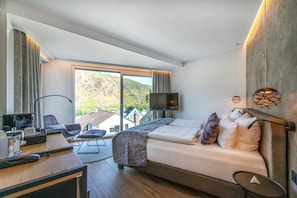 A modern hotel room with a large bed, a desk, a chair, a TV, and a balcony with a view of greenery.