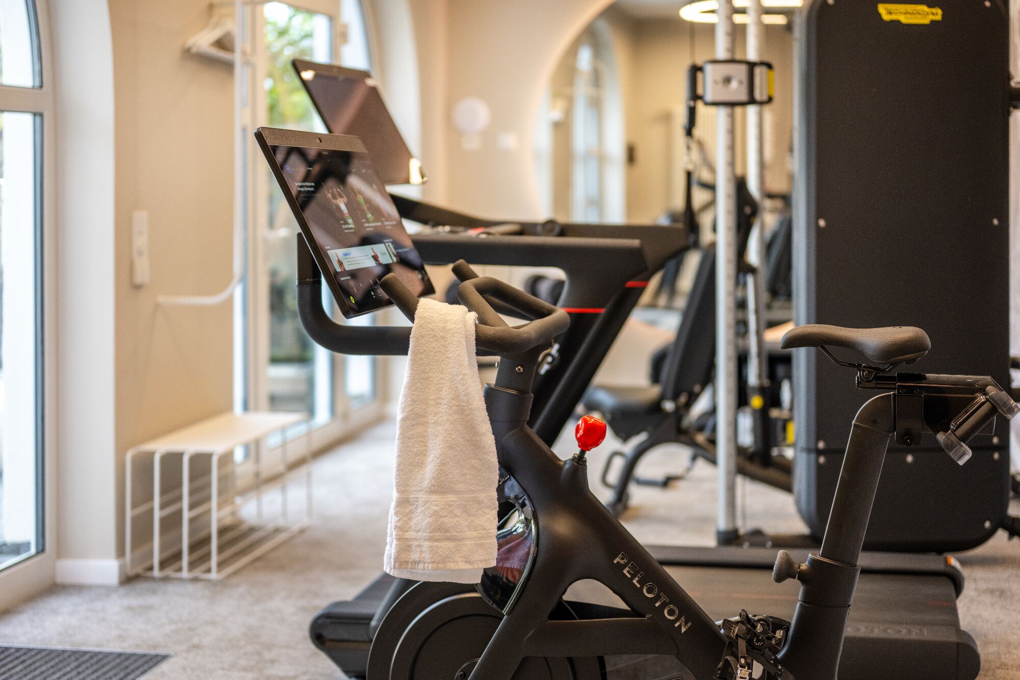 An exercise bike with a digital display, a towel, and a treadmill in the background.