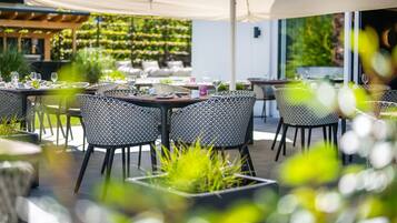 Un área de comedor al aire libre con mesas y sillas, rodeada de follaje y un paraguas grande.