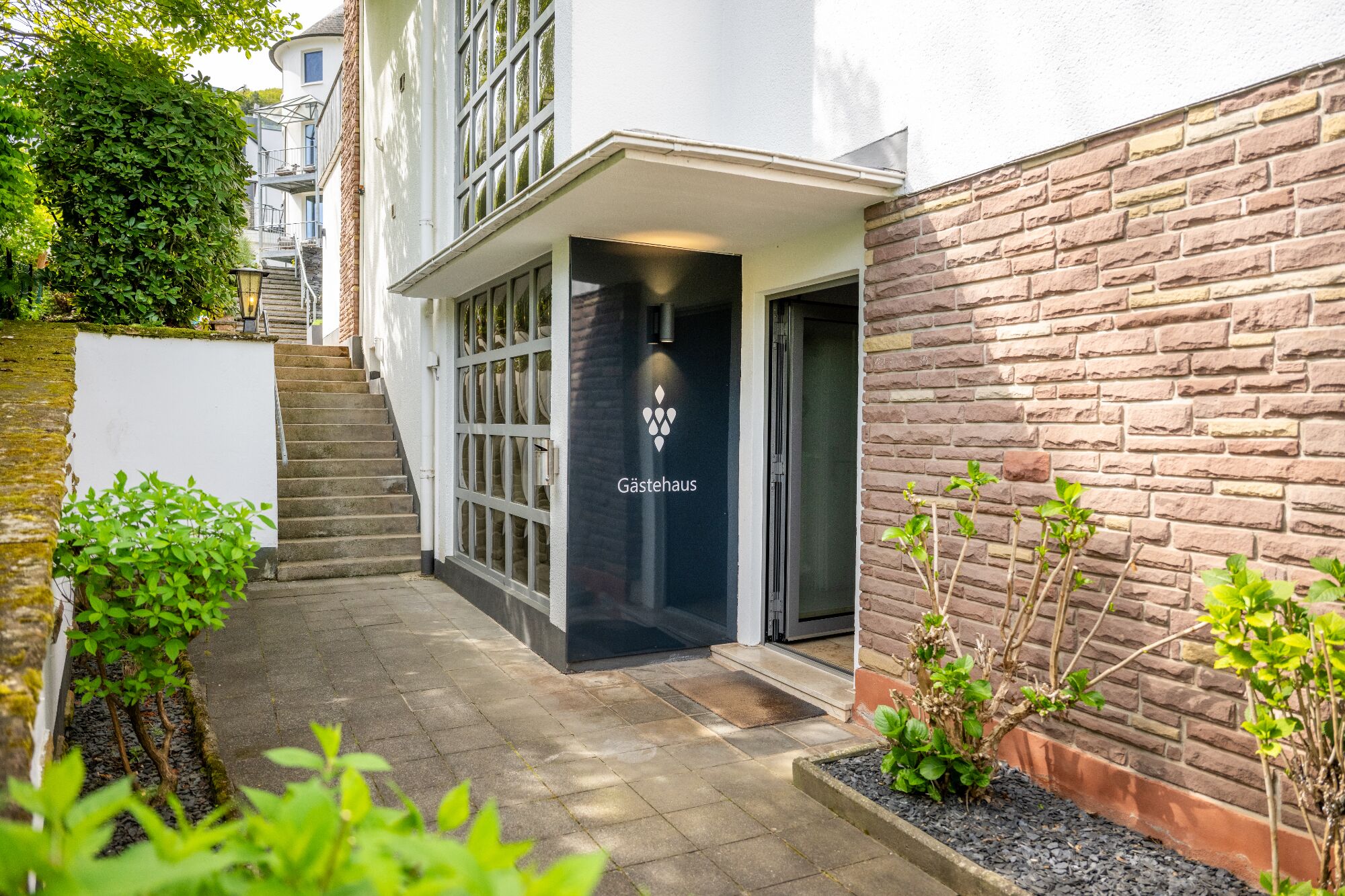 Un bâtiment moderne avec une porte d’entrée noire portant l’inscription « Gästehaus », un mur de briques et un petit jardin agrémenté de plantes.
