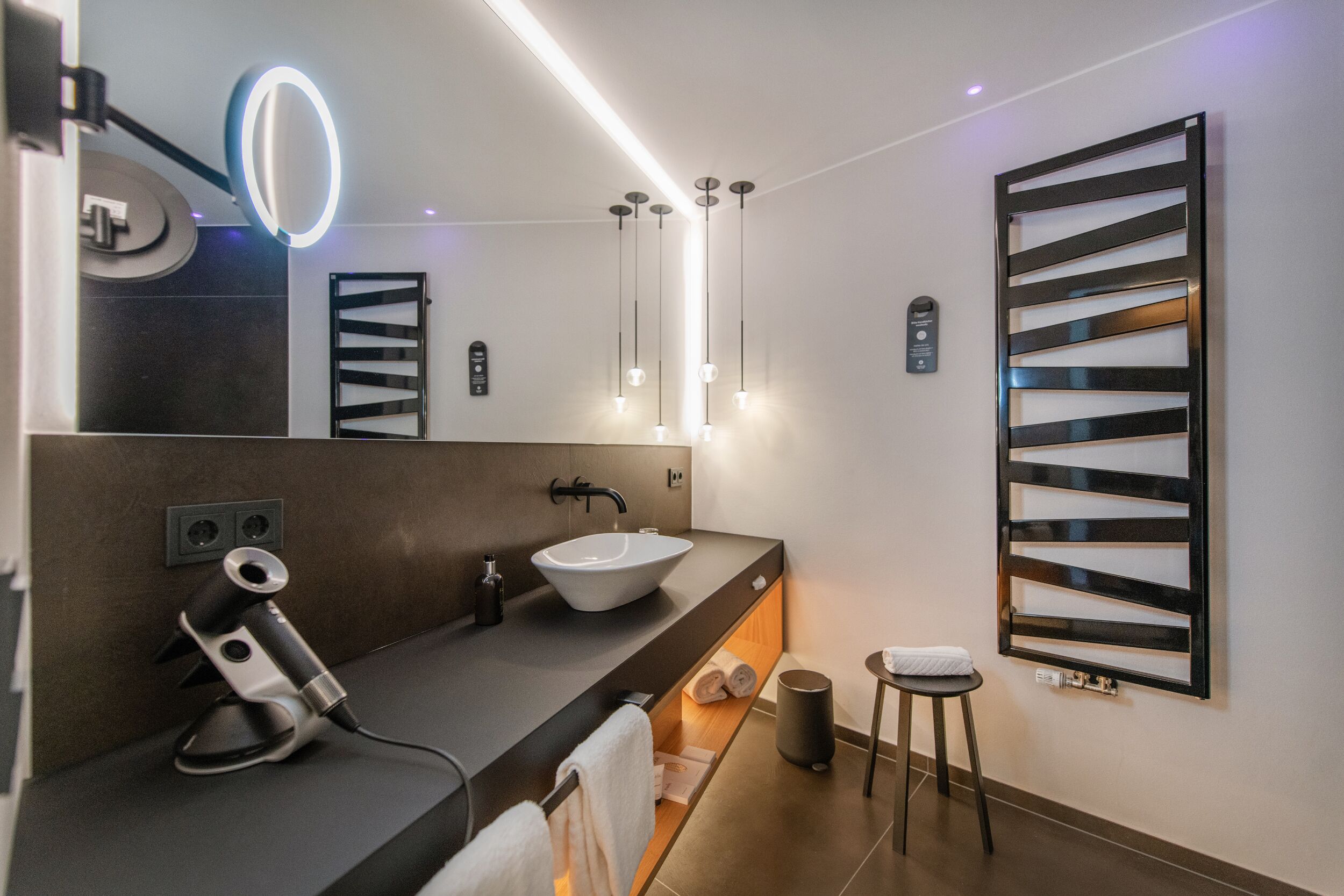 Ein modernes Badezimmer mit dunkler Ablage, einem weißen Waschbecken, einem Spiegel mit Beleuchtung, einem Handtuchhalter und einem kleinen Hocker.