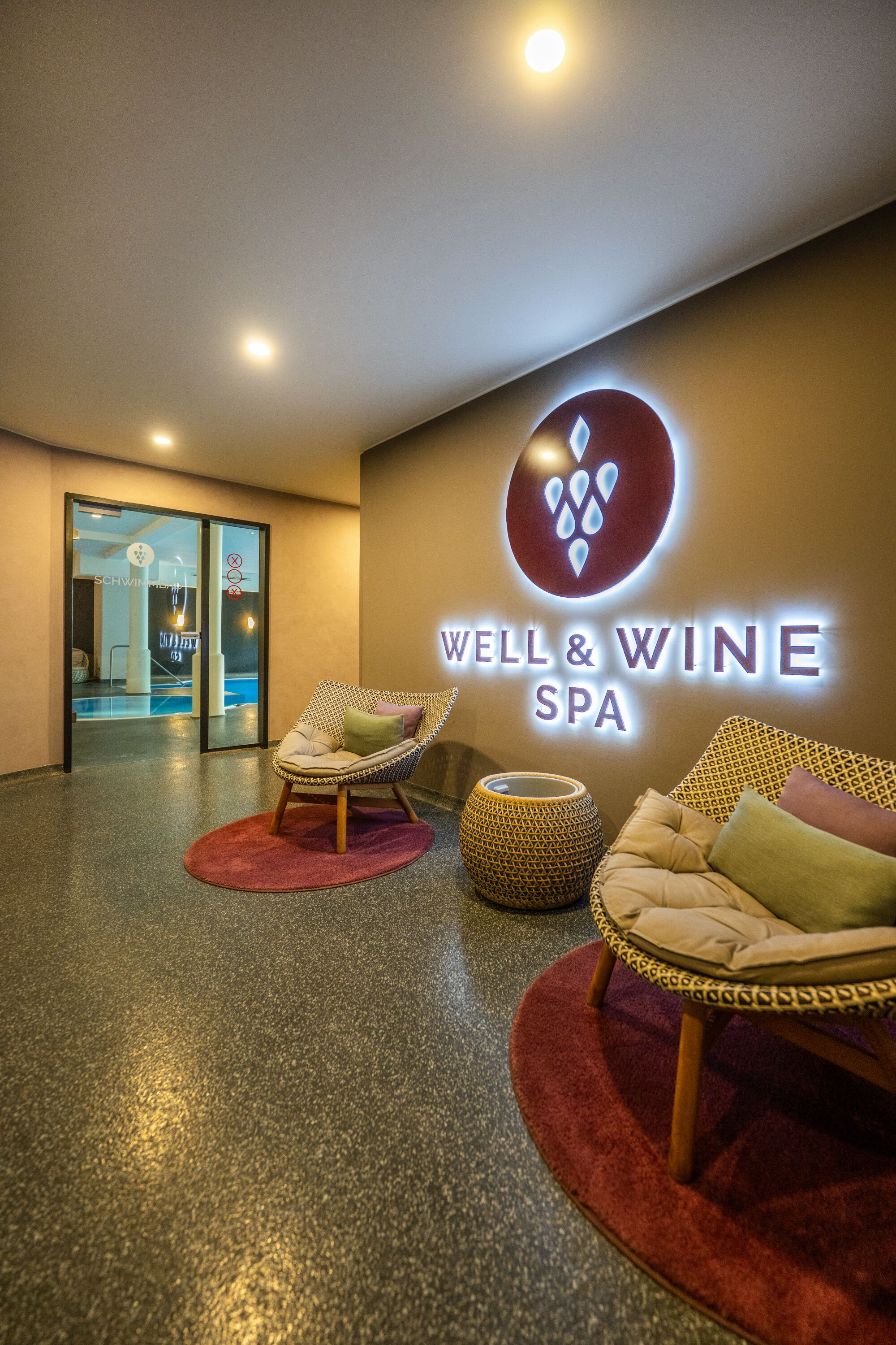 Un espace de bien-être avec une enseigne indiquant « SPA WELL & WINE », deux fauteuils en osier avec des coussins, un tapis rond et un sol texturé.