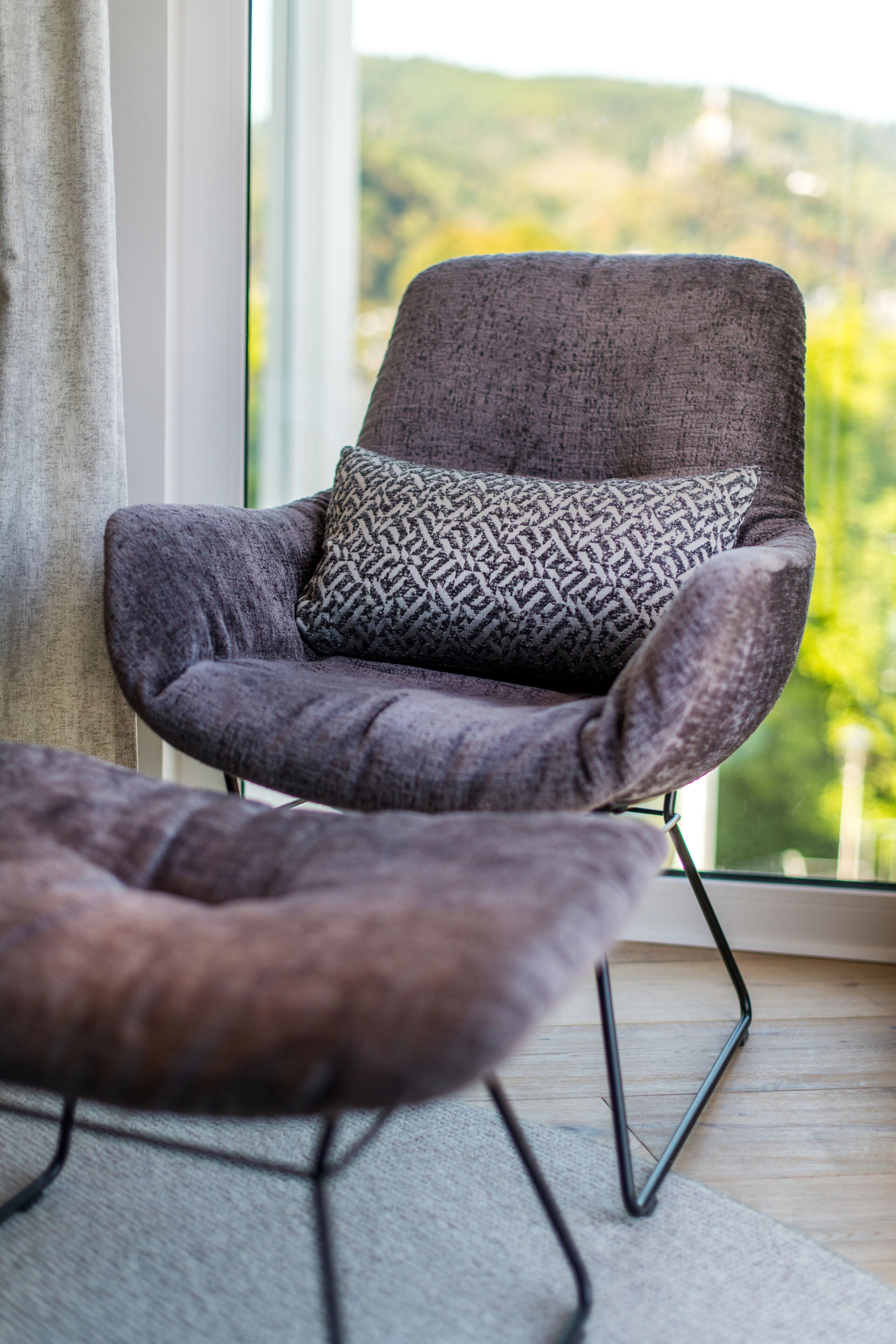 A modern armchair with a patterned cushion, positioned near a window with a view of greenery.