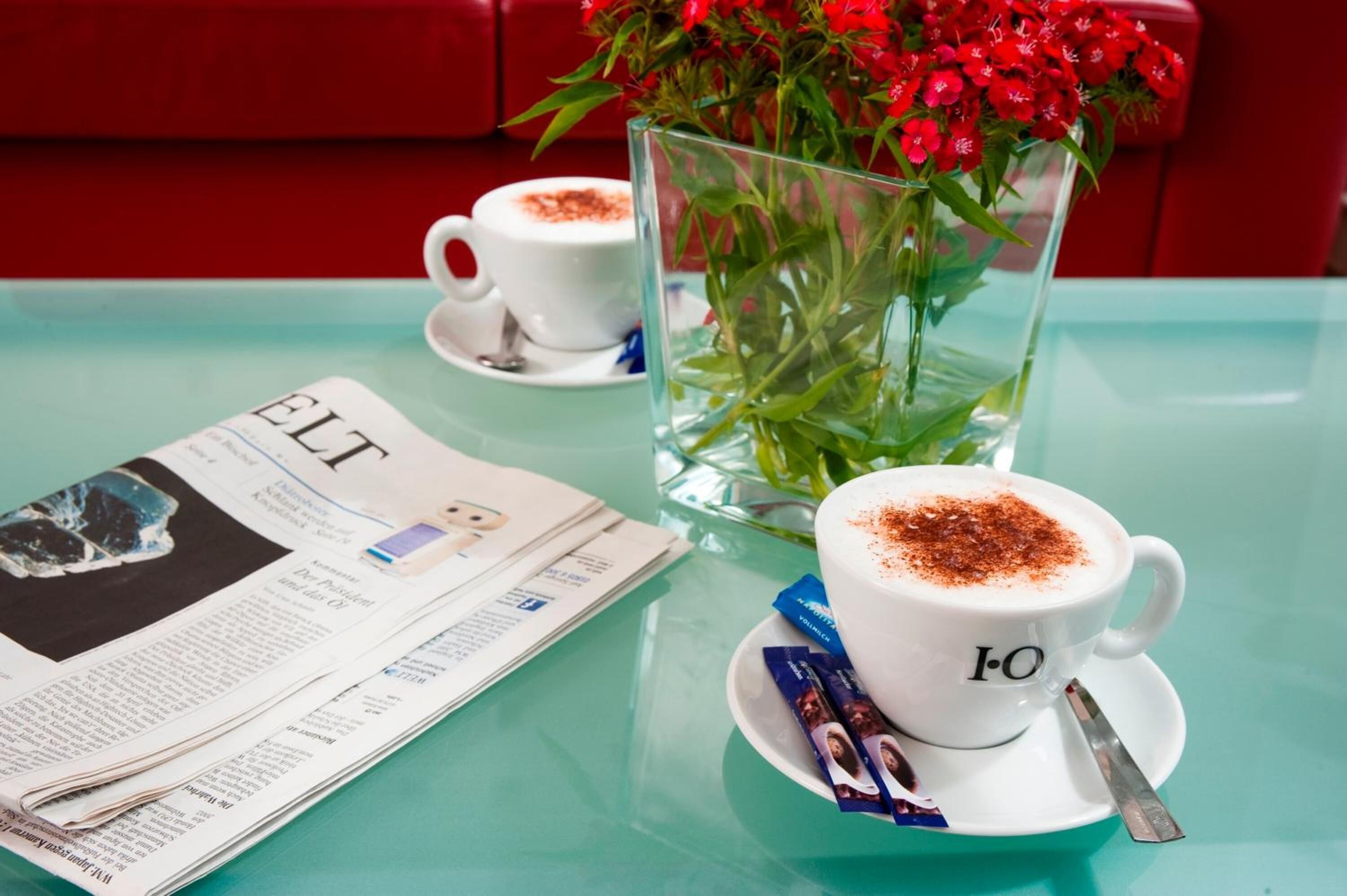 A table with a newspaper, a cup of coffee with a spoon, a glass vase with flowers, and a red couch in the background.