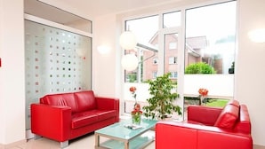 A modern living room with two red sofas, a glass coffee table, and a large window offering a view of a residential area. - Hotel Aquamarin (Papenburg)