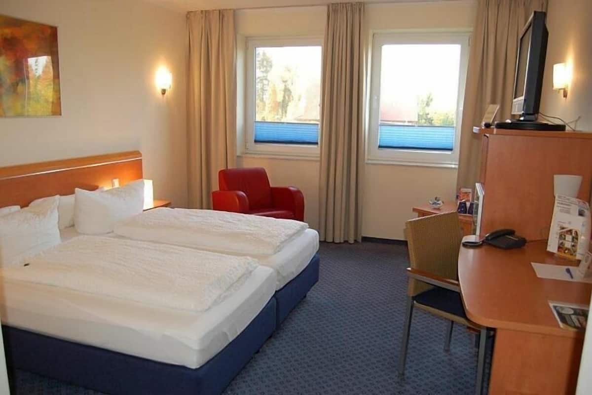 Una habitación de hotel con una cama doble, un escritorio con silla, un televisor y dos ventanas con cortinas.