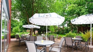 An outdoor patio with round tables and chairs, large umbrellas, and lush greenery in the background. - Hotel Aquamarin (Papenburg)