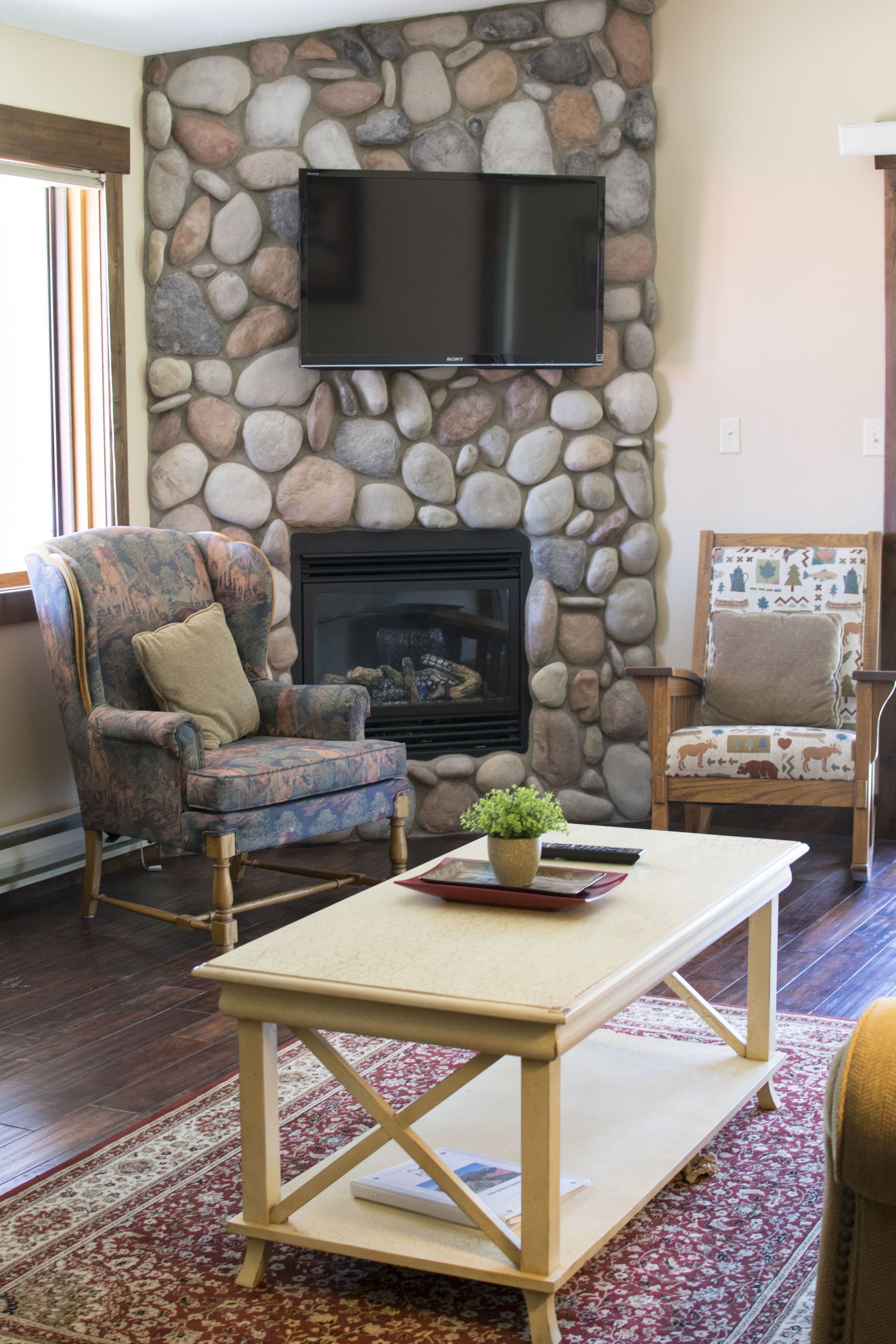 Una sala de estar con chimenea de piedra, un televisor de pantalla plana montado encima, dos sillones con estampado, una mesa de centro y un tapete con motivos rojos y blancos.