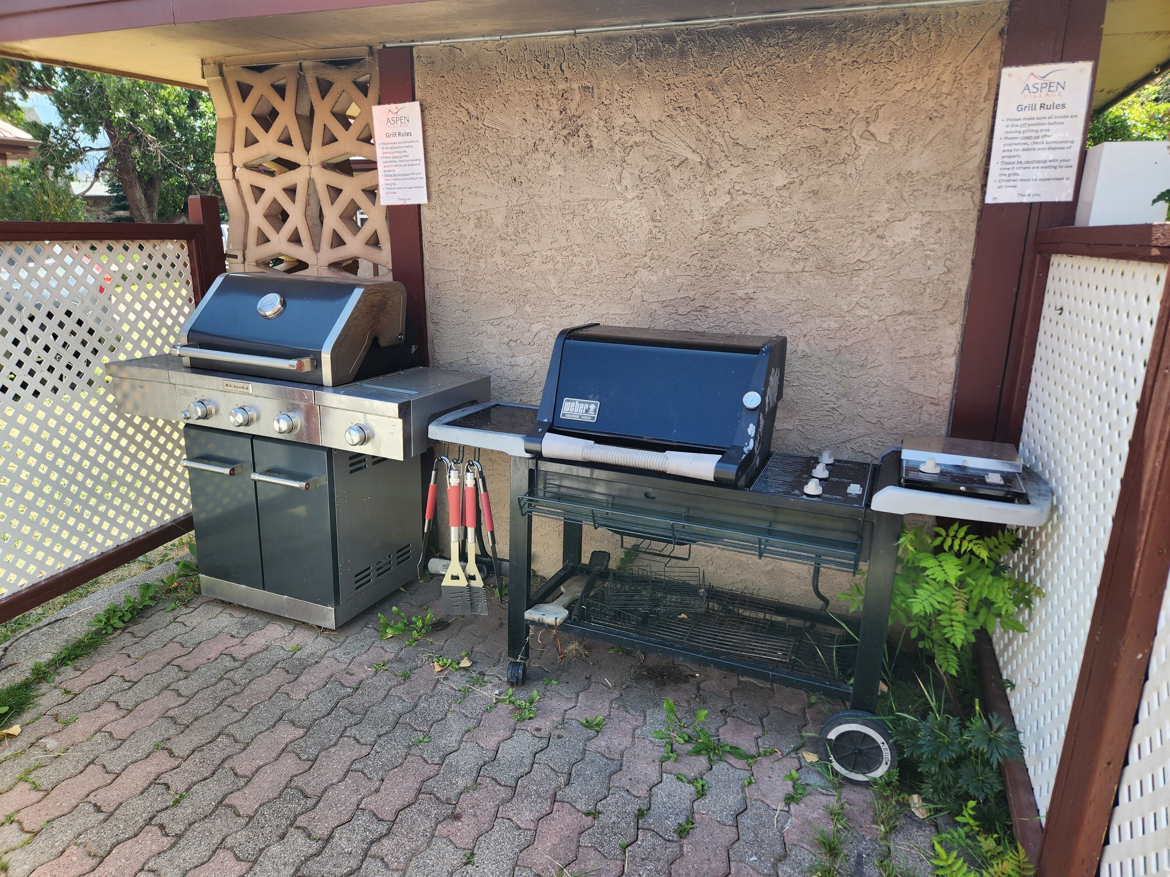 Un espace barbecue extérieur équipé d’un barbecue de marque Weber et d’un barbecue portable, entouré d’une terrasse en briques et d’une clôture en treillis.