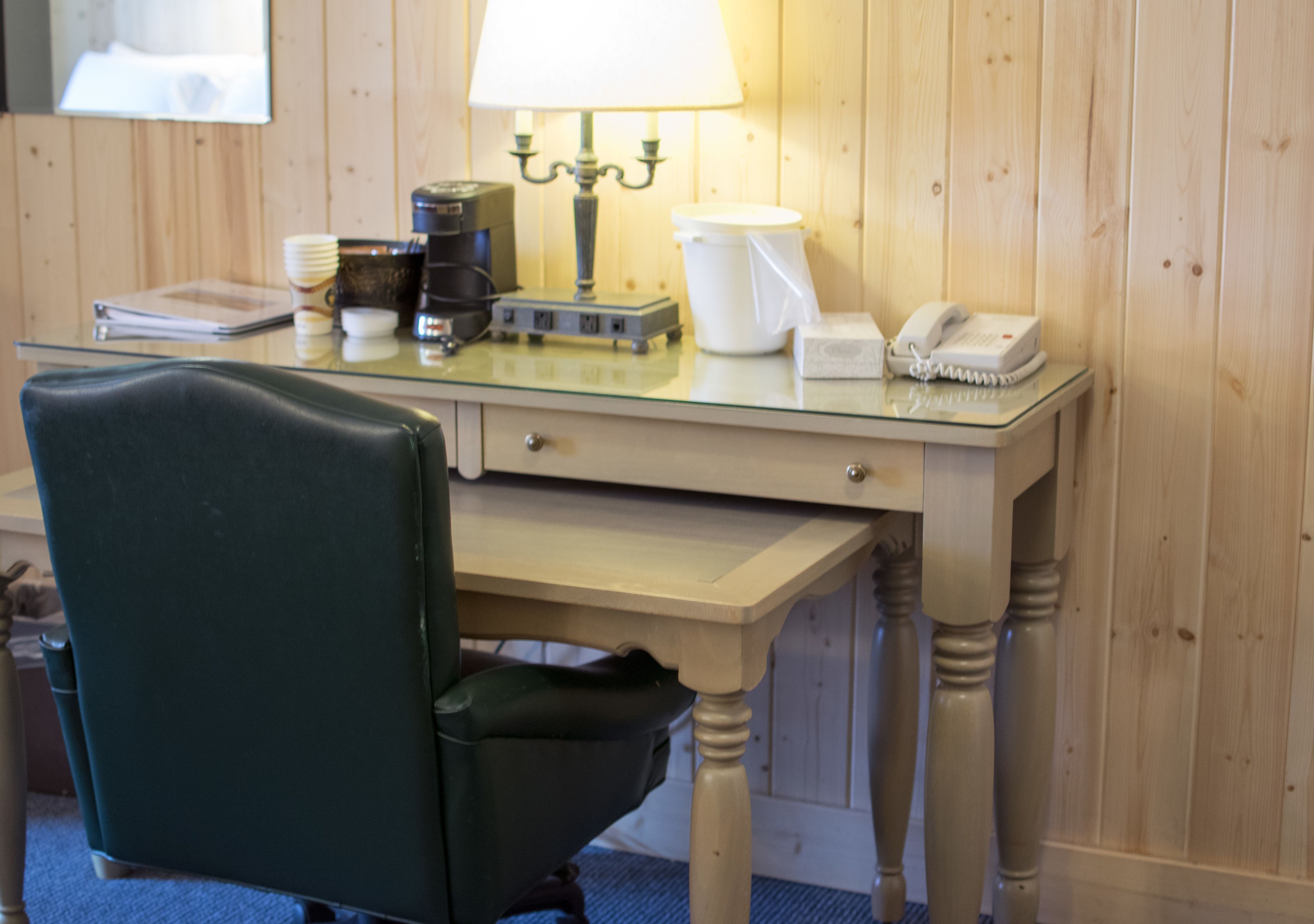 A hotel room with a wooden paneled wall, a glass desk with a lamp, a telephone, a coffee maker, and a chair.
