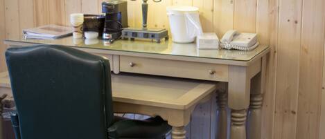 A hotel room with a wooden paneled wall, a glass desk with a lamp, a telephone, a coffee maker, and a chair.