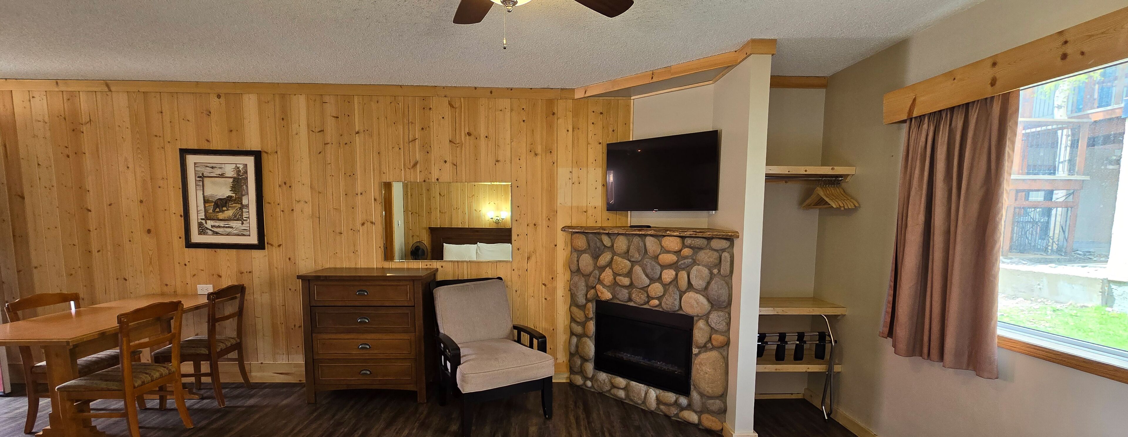 A room with wooden panel walls, a stone fireplace, a television mounted above it, a wooden dining table with chairs, a dresser, and a window with curtains.