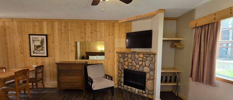 A room with wooden panel walls, a stone fireplace, a television mounted above it, a wooden dining table with chairs, a dresser, and a window with curtains.