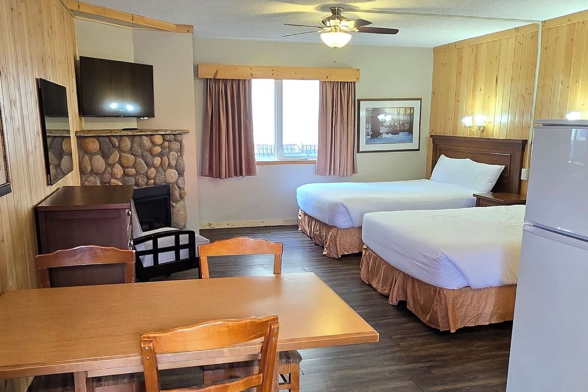A room with a wooden dining table, two beds, a TV, and a ceiling fan.