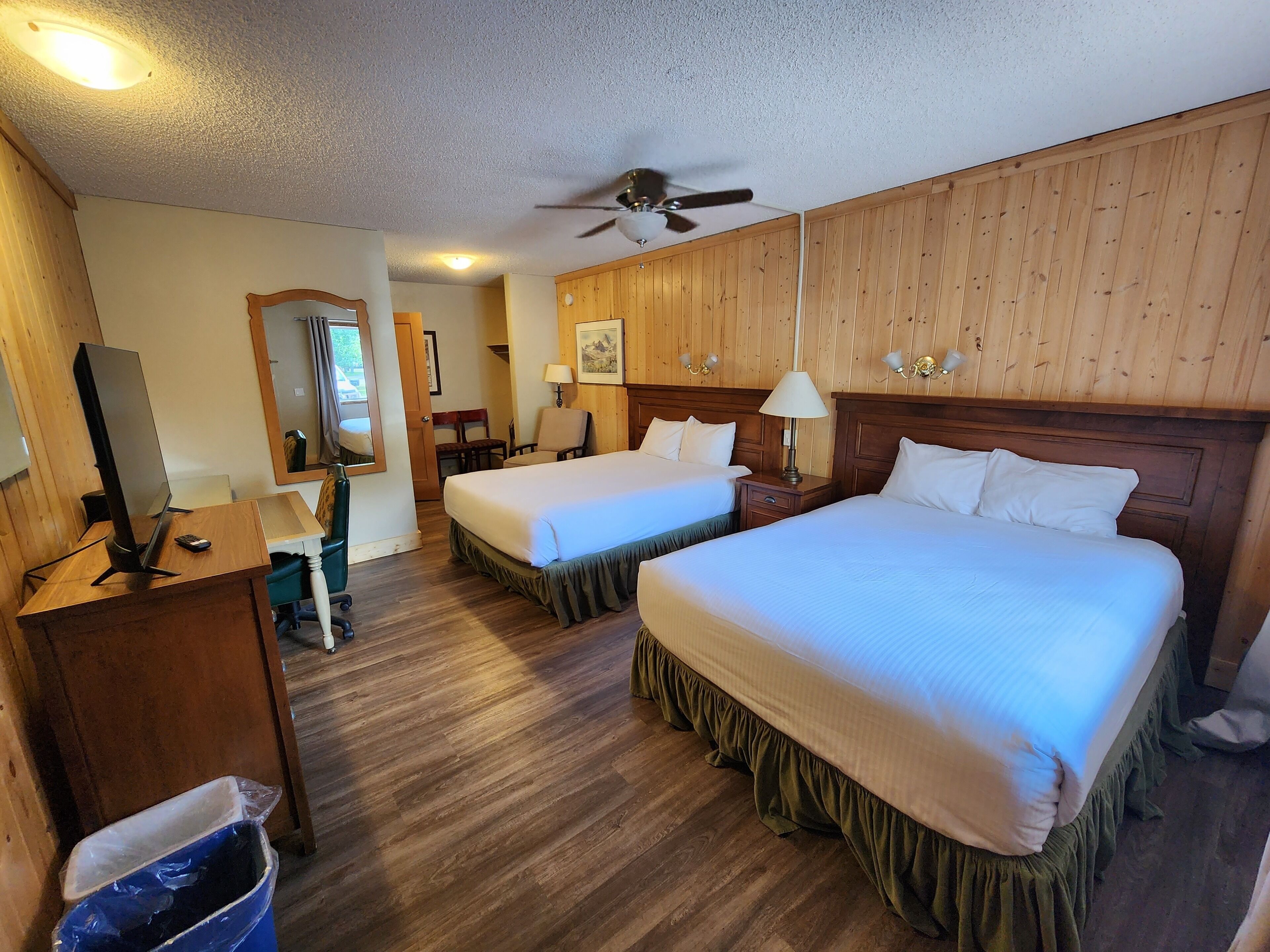 Habitación de hotel con dos camas, un escritorio con ordenador, televisor, ventilador de techo y paneles de madera en las paredes.
