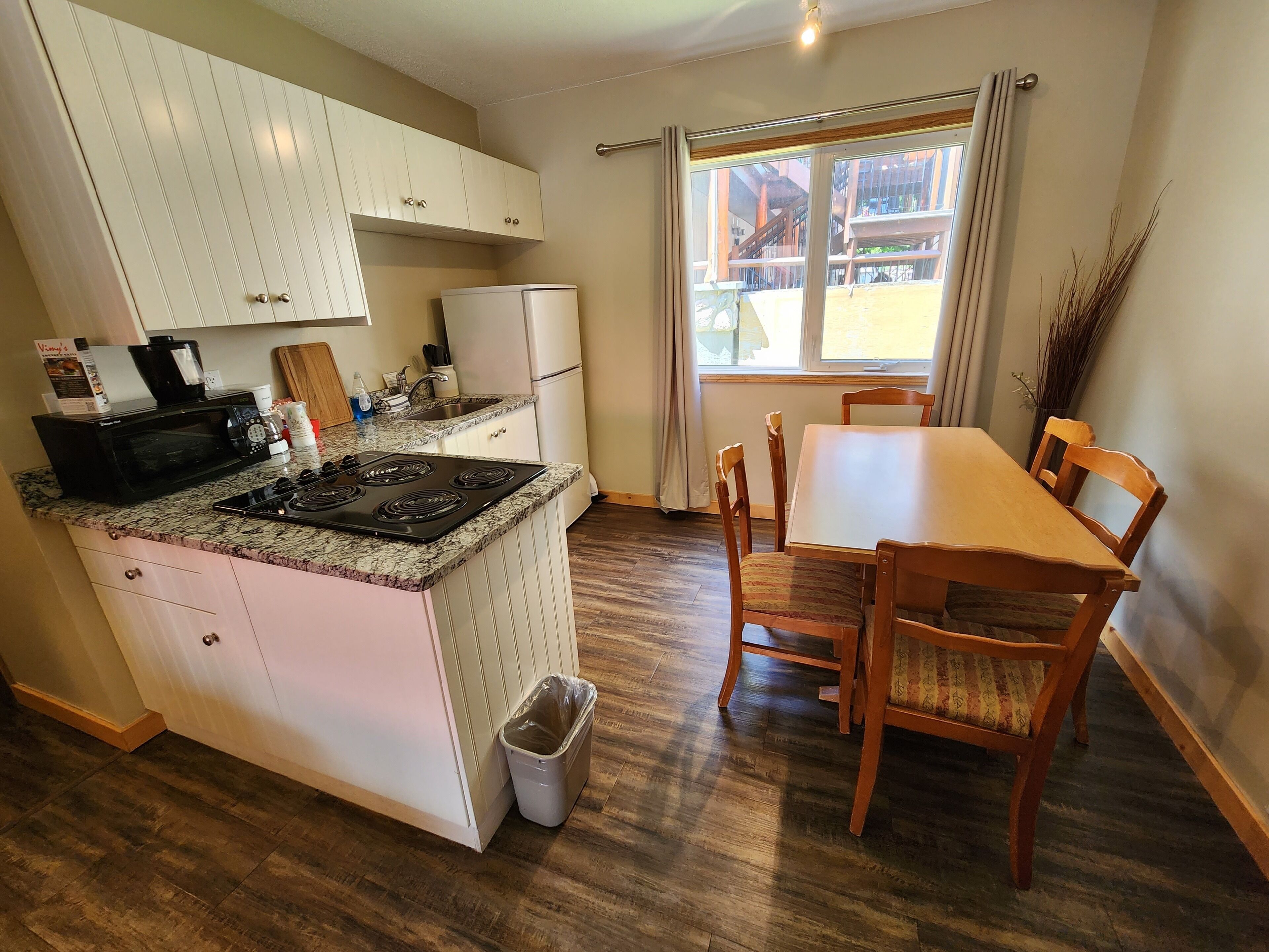Een kleine keuken met een fornuis, magnetron en koelkast, een eettafel met stoelen en een uitzicht door een raam.