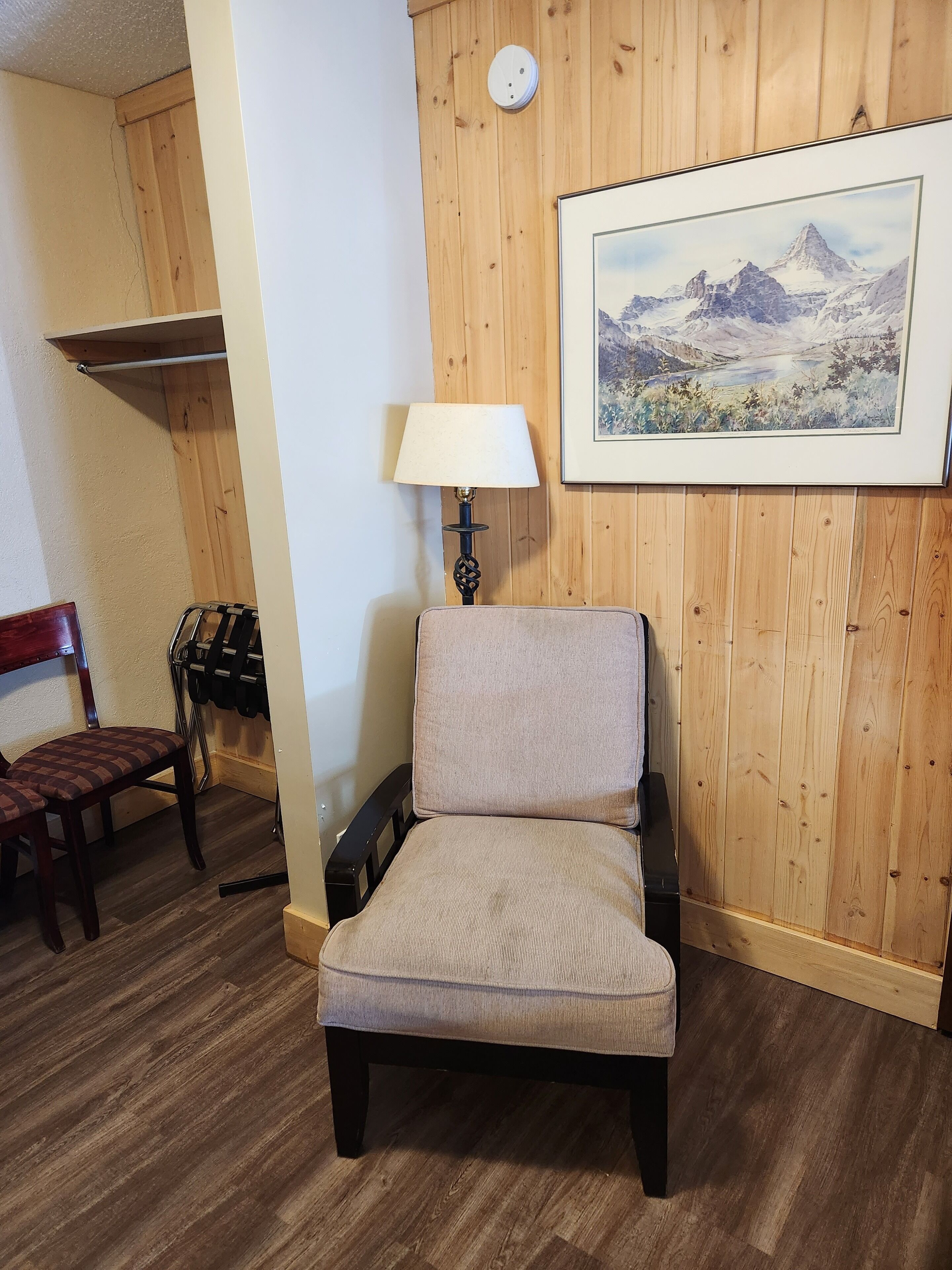 A room with a wooden paneled wall, a framed landscape painting, a chair with a cushion, a lamp, and a wooden floor.