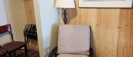 A room with a wooden paneled wall, a framed landscape painting, a chair with a cushion, a lamp, and a wooden floor.