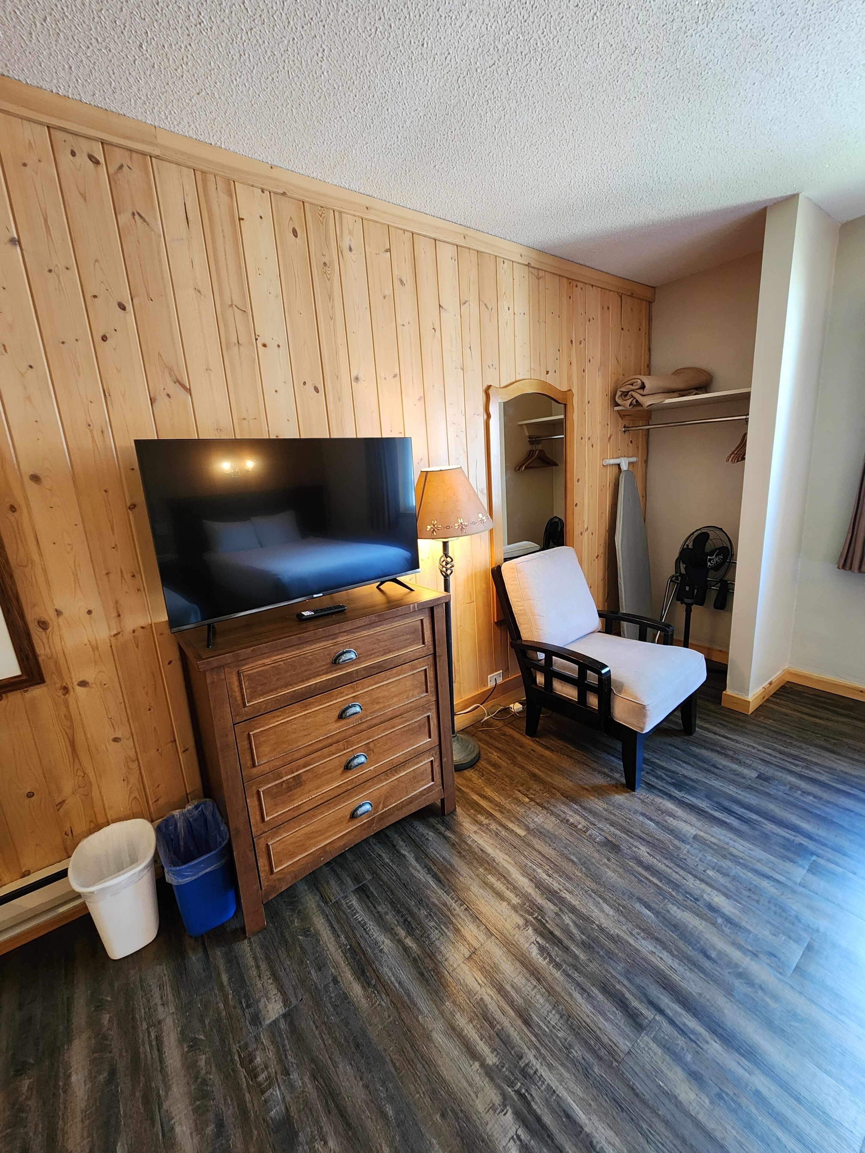 A room with wooden panel walls, a television on a wooden cabinet, a chair, a small trash bin, and a fan.