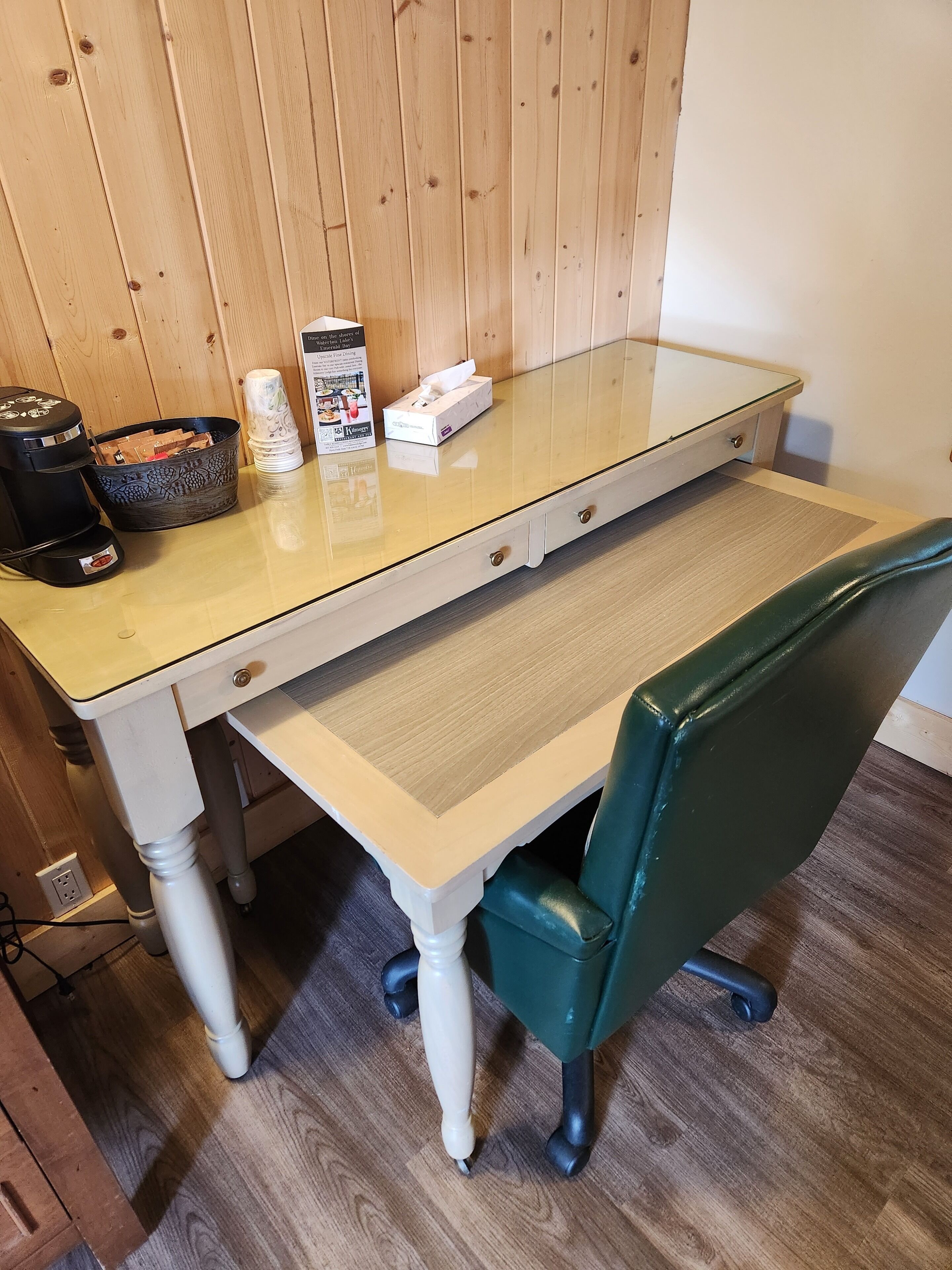 Een kamer met houten lambrisering, een tafel met glazen blad, een zwarte koffiezetapparaat, een mand en een zakdoos.