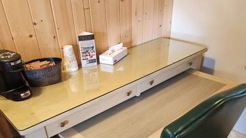 Een kamer met houten lambrisering, een tafel met glazen blad, een zwarte koffiezetapparaat, een mand en een zakdoos.