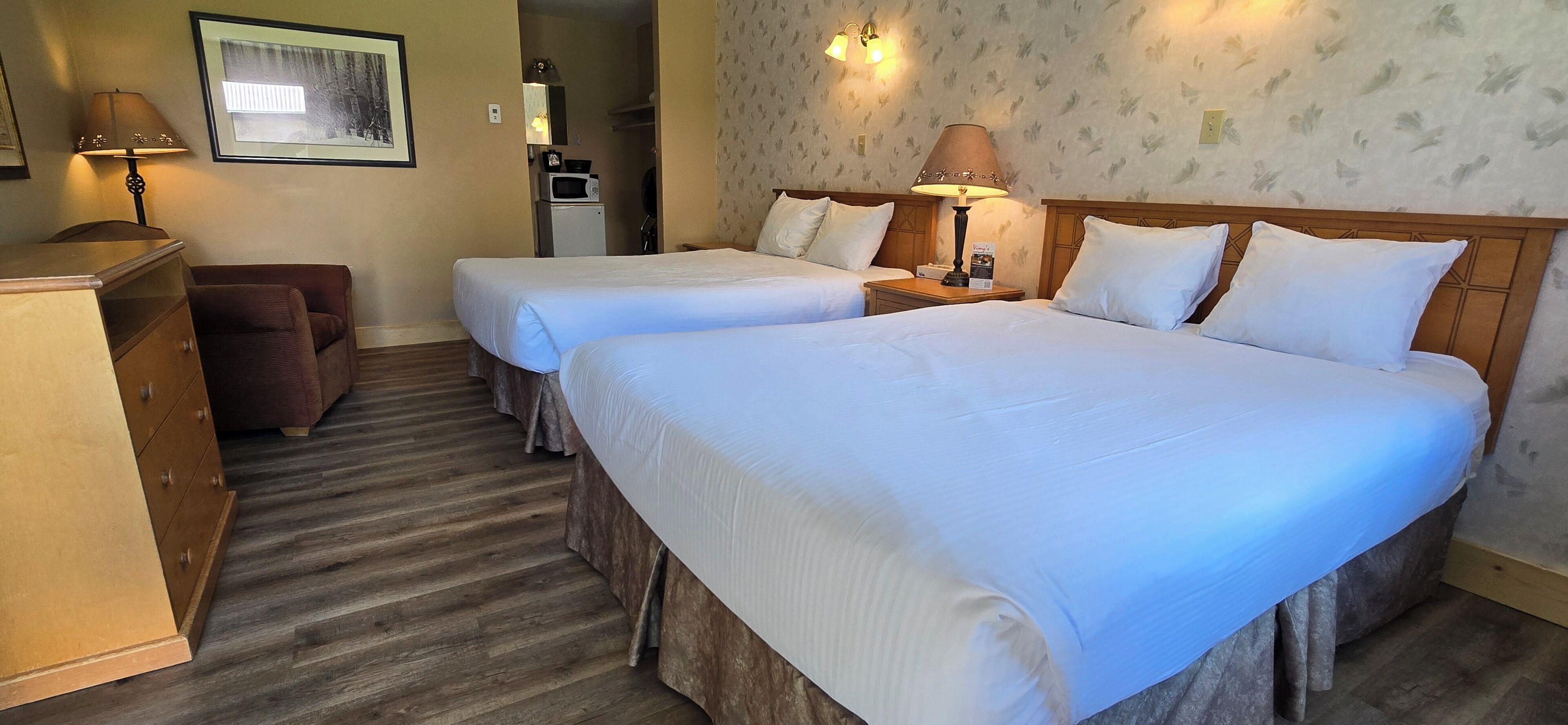 Ein Hotelzimmer mit zwei Betten, einem Holzkoffer, einem Schreibtisch, einer Lampe, einer Mikrowelle und einem gerahmten Bild an der Wand.