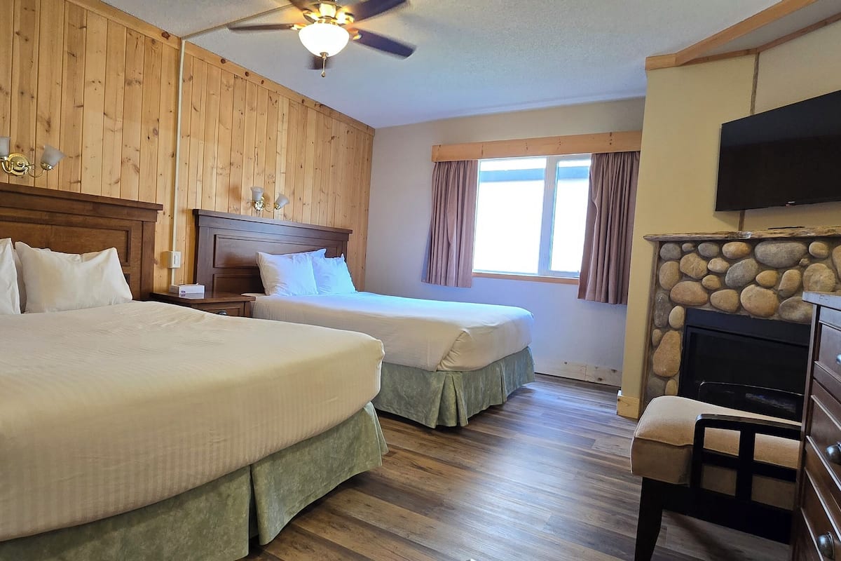 A hotel room with two beds, wooden paneling, a stone fireplace, a television, and a ceiling fan.