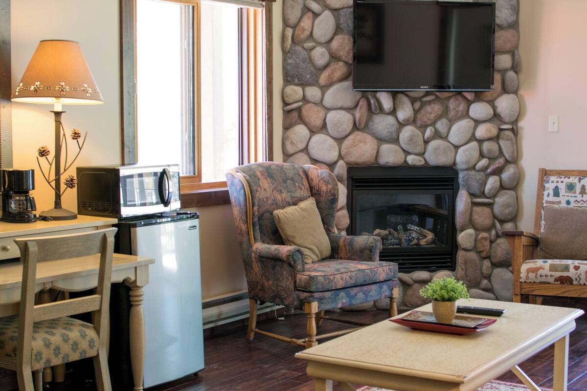 A cozy living room with a stone fireplace, a comfortable armchair, a dining table, and a television mounted on the wall.