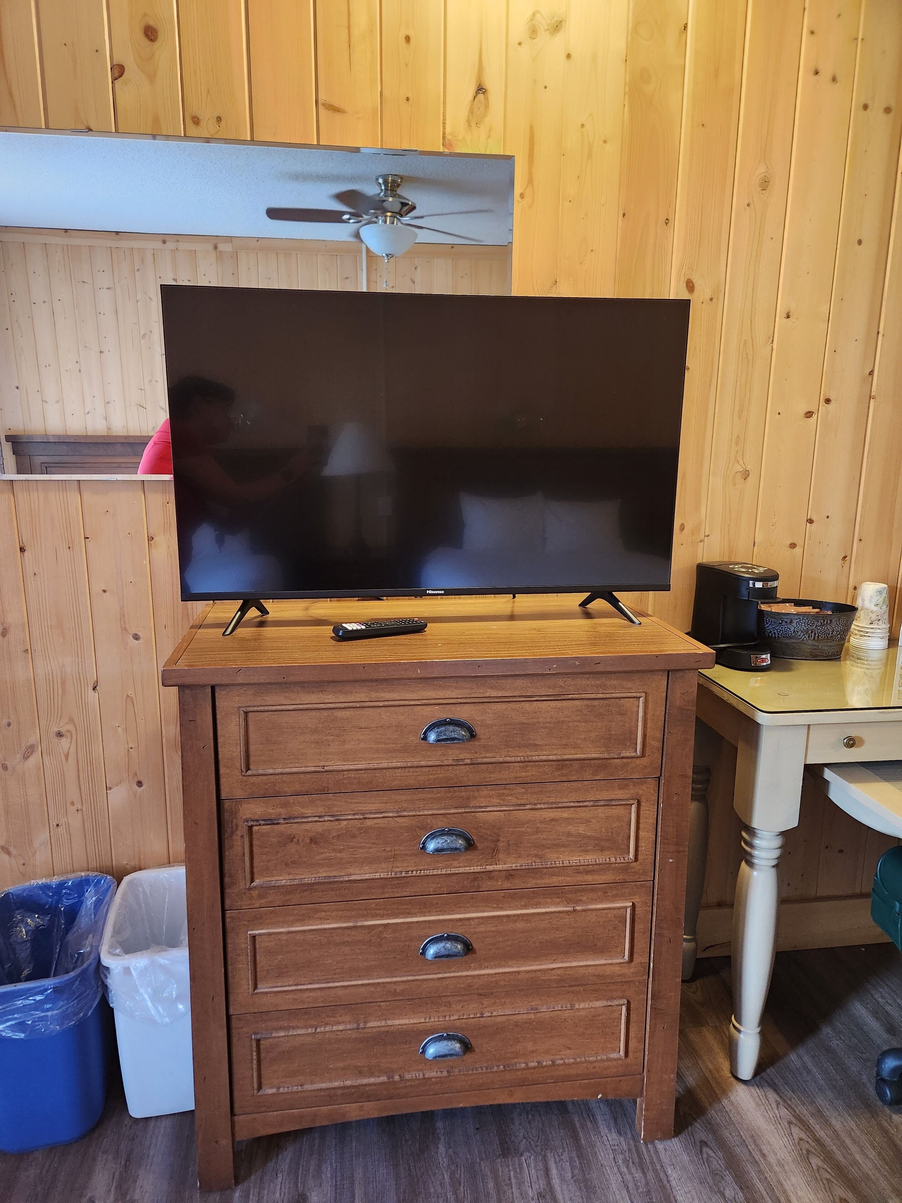 Een houten kast met een flatscreen televisie erop, een plafondventilator erboven en een klein tafeltje met een koffiezetapparaat en een kom.