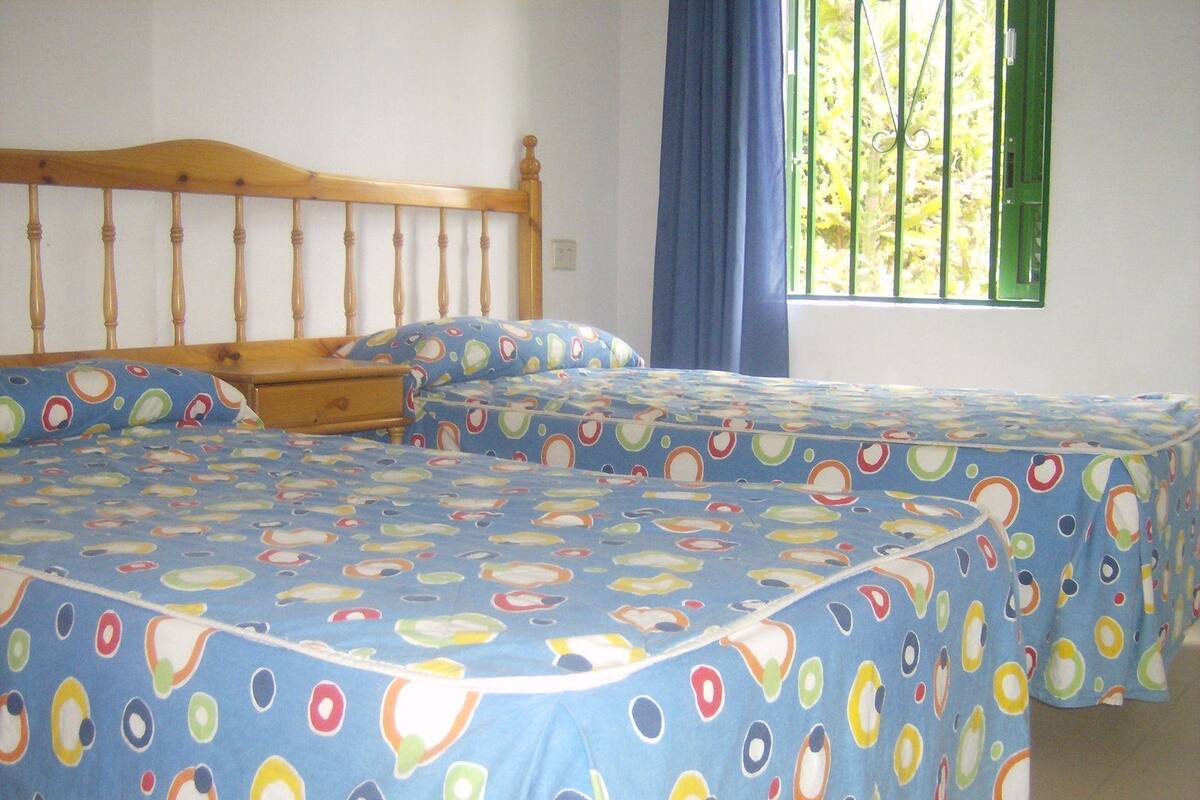 A room with two beds, each with a blue bedspread featuring colorful circular patterns. There is a wooden headboard, a window with green blinds, and a blue curtain.