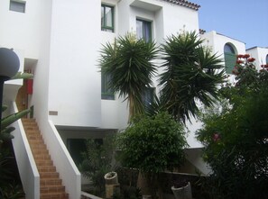 A white building with a staircase leading to a door, surrounded by various plants and flowers.