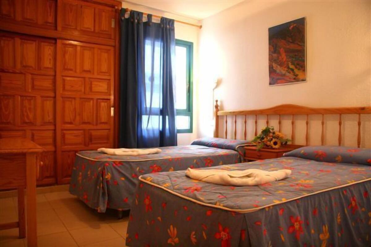 A room with two beds, a wooden wardrobe, a window with curtains, and a painting on the wall.
