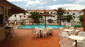 A poolside area with white plastic chairs and tables, a thatched umbrella, and a building with a tiled roof in the background.