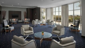 A modern hotel lobby with a central round table, comfortable seating, a flat-screen TV, and large windows offering a view of the outdoors. - Courtyard Phoenix Chandler/Fashion Center (Chandler)