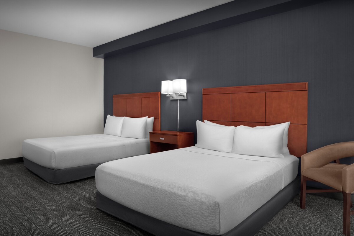 A hotel room with two beds, a nightstand, a lamp, and a chair.