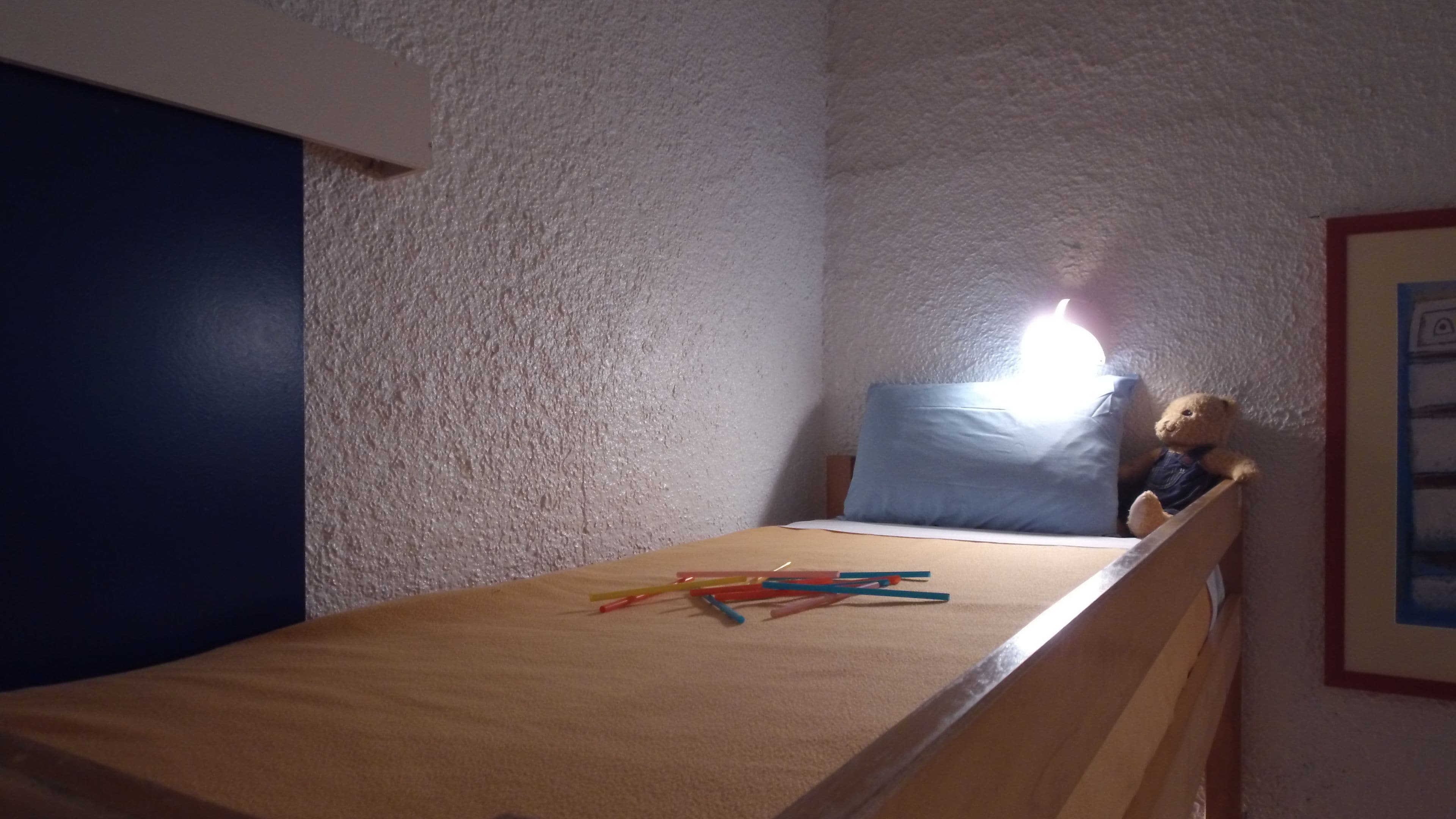 Une petite chambre avec un lit simple, une tête de lit en bois, un oreiller bleu et un ours en peluche.