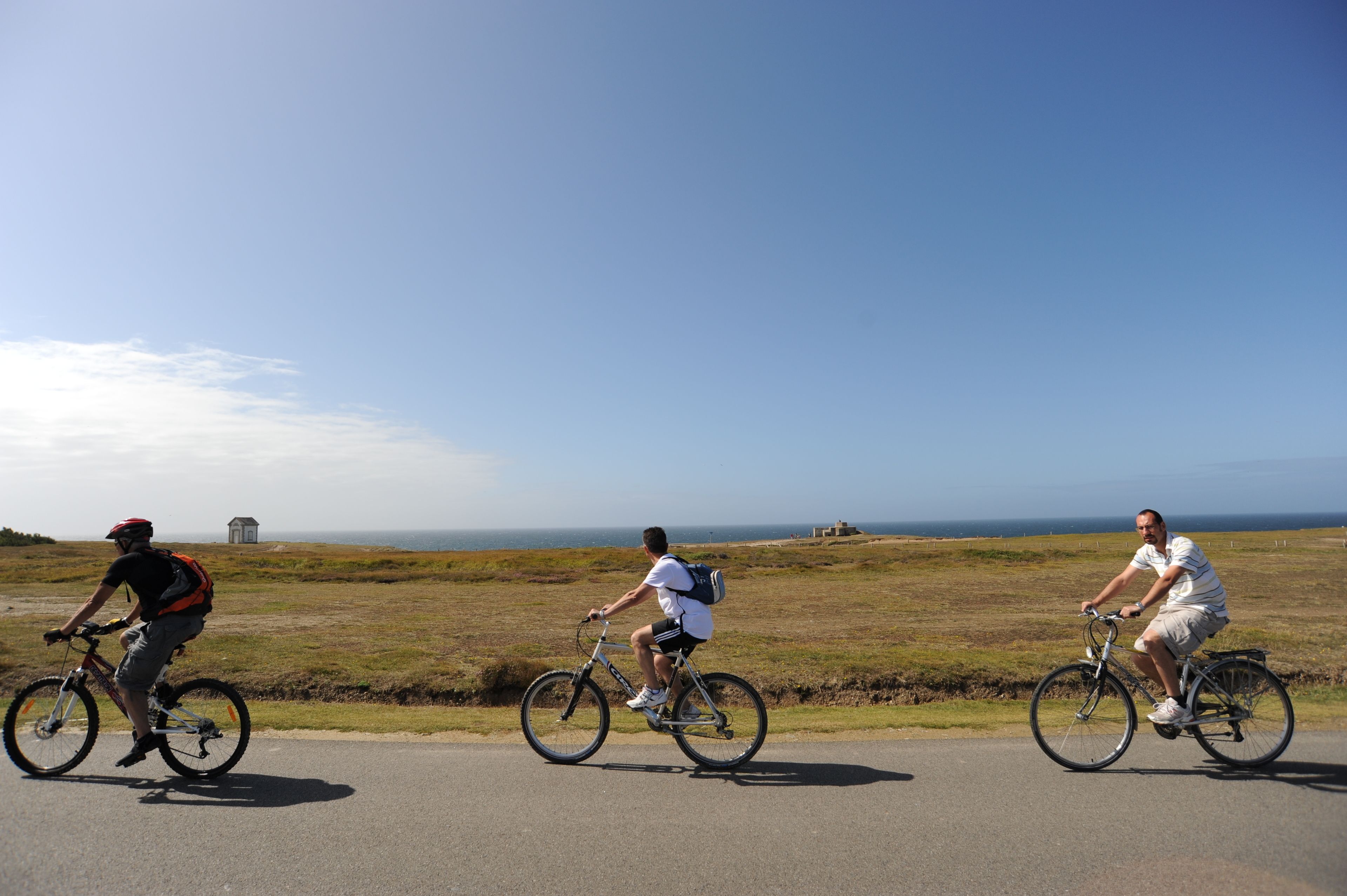 Three cyclists on a road with a grassy field and ocean in the background.