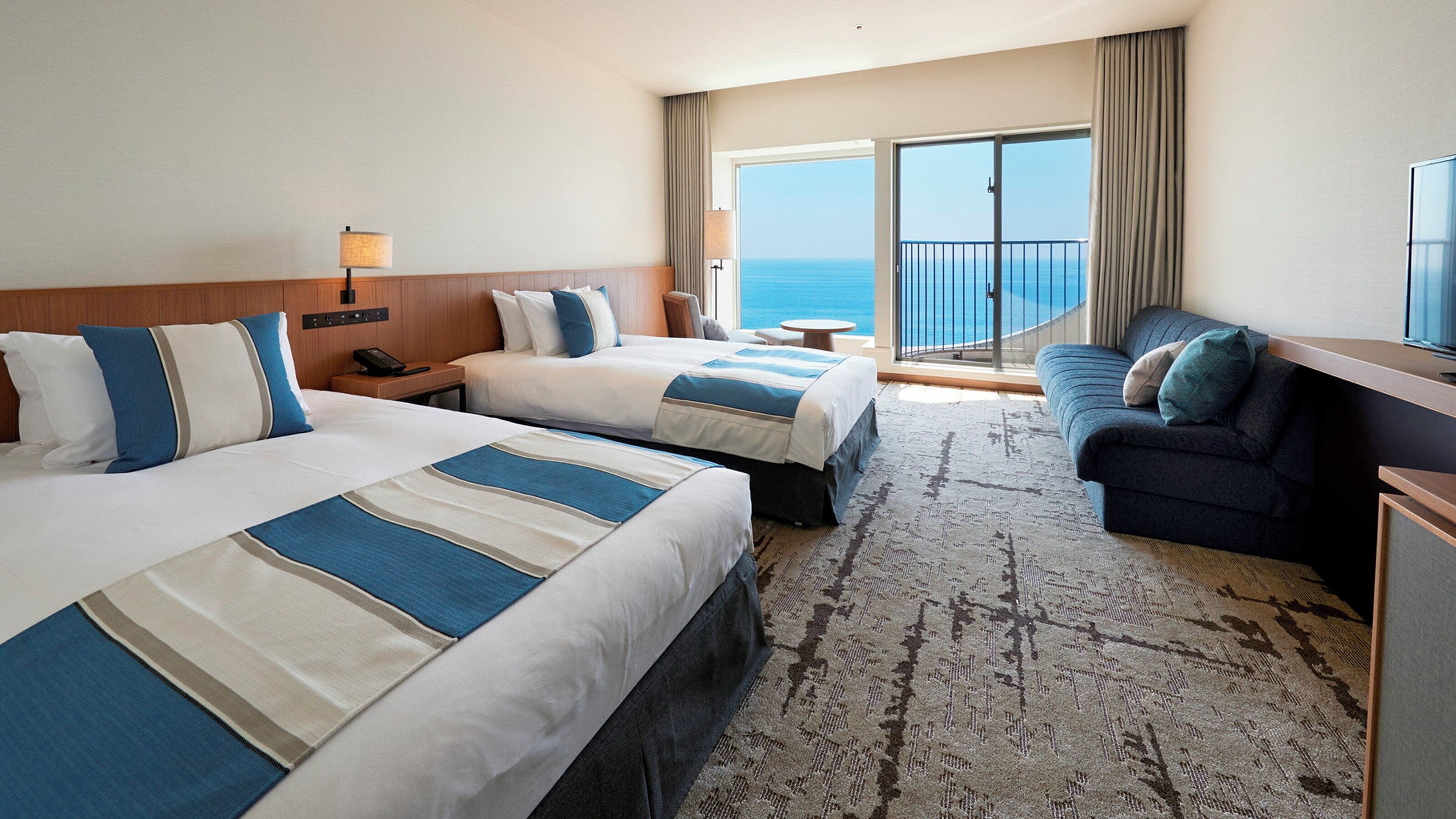 A hotel room with two beds, a sofa, a desk, and a balcony with a view of the ocean.