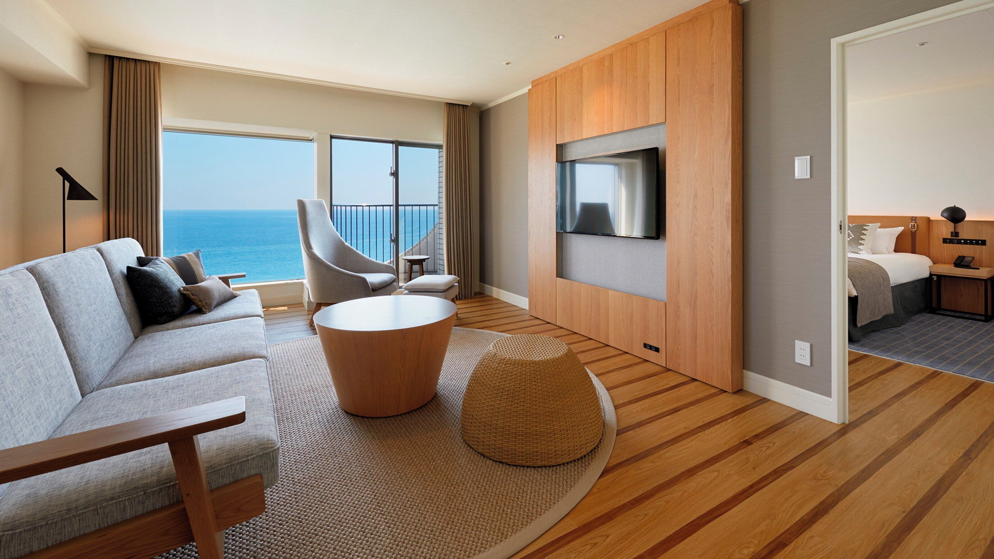 Una habitación de hotel moderna con sofá, una mesa de centro redonda, un televisor montado en la pared y un balcón con vista al mar.