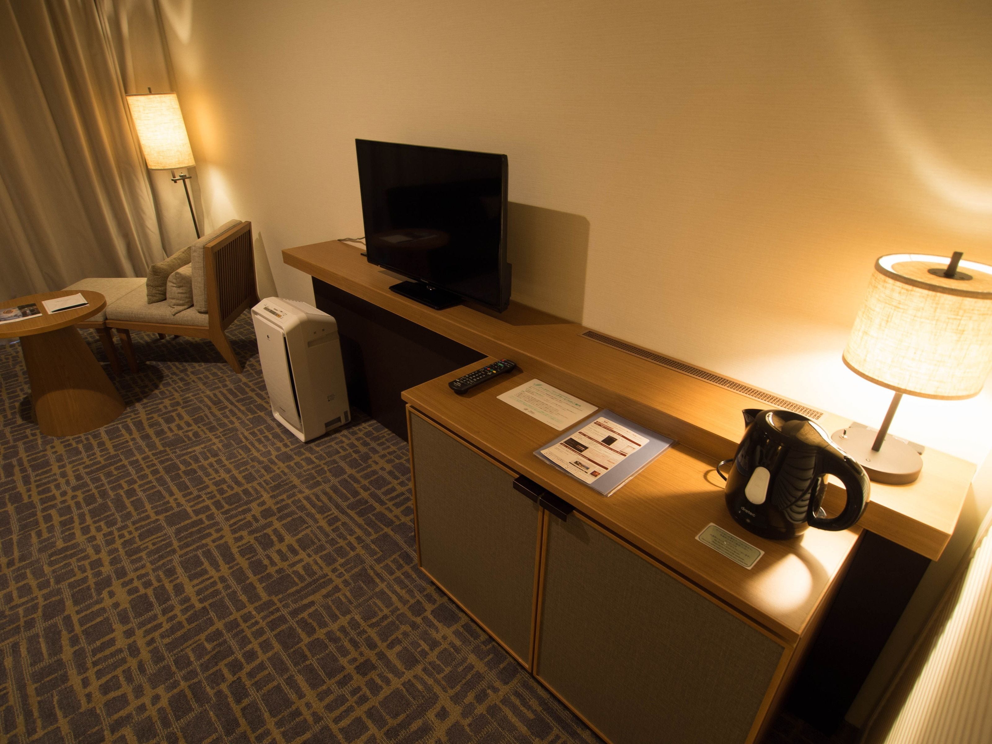 A hotel room with a desk, a television, a lamp, a chair, a coffee maker, and a small refrigerator.