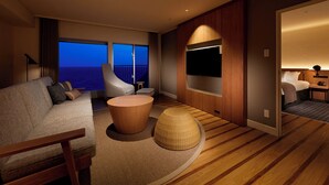 A modern hotel room with a large window offering a view of the ocean, a sofa, a round wooden coffee table, and a television mounted on the wall. - Oiso Prince Hotel (Oiso)