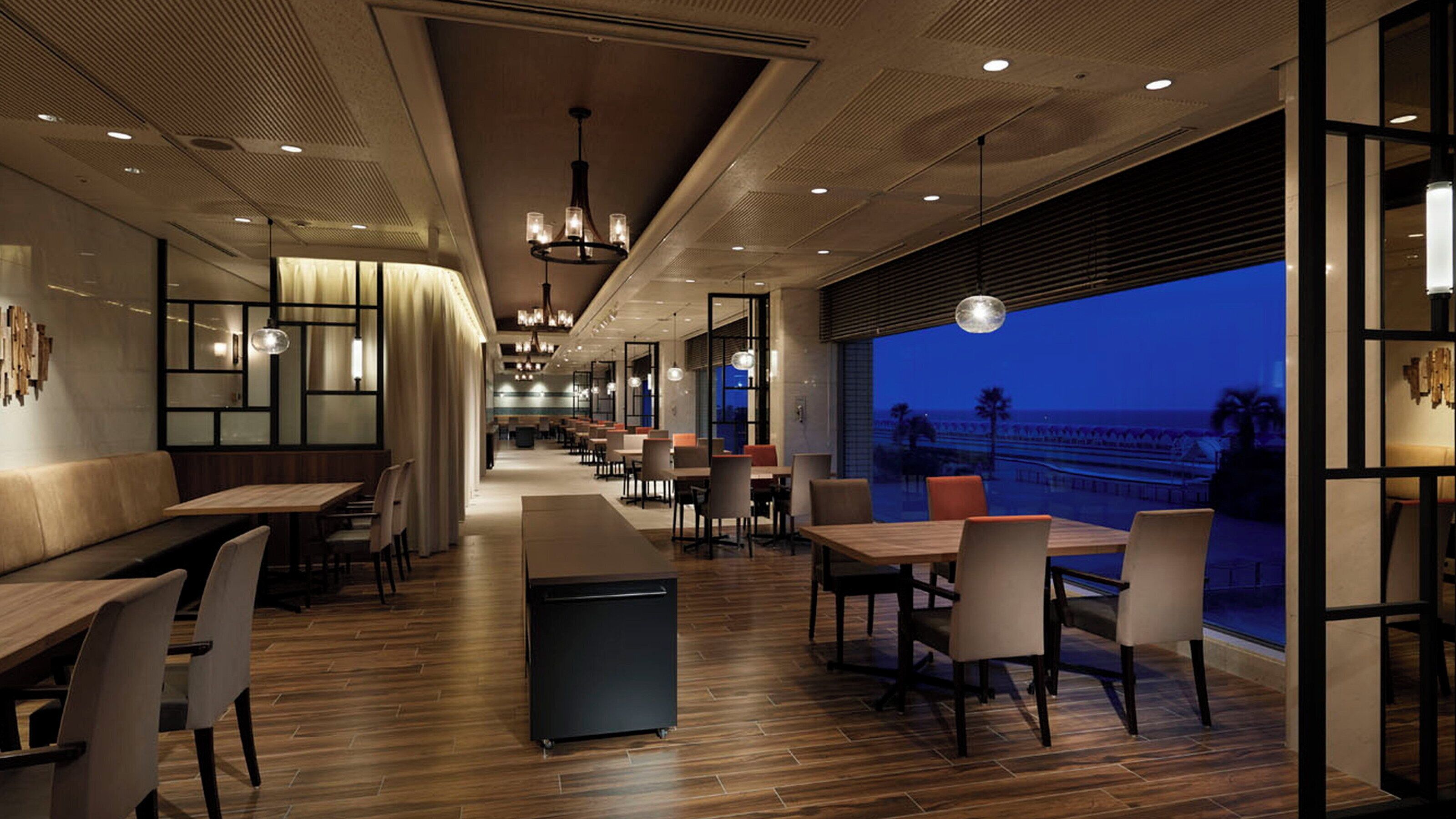 A modern restaurant with wooden flooring, multiple seating areas including tables and booths, and large windows offering a view of the sea.
