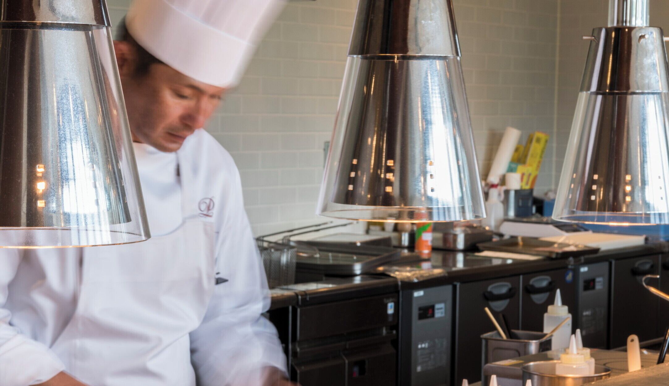 Un chef vêtu d’une blouse blanche et coiffé d’un chapeau de chef, travaillant derrière un plan de travail en acier inoxydable, éclairé par des lampes suspendues, et entouré de divers appareils de cuisine.