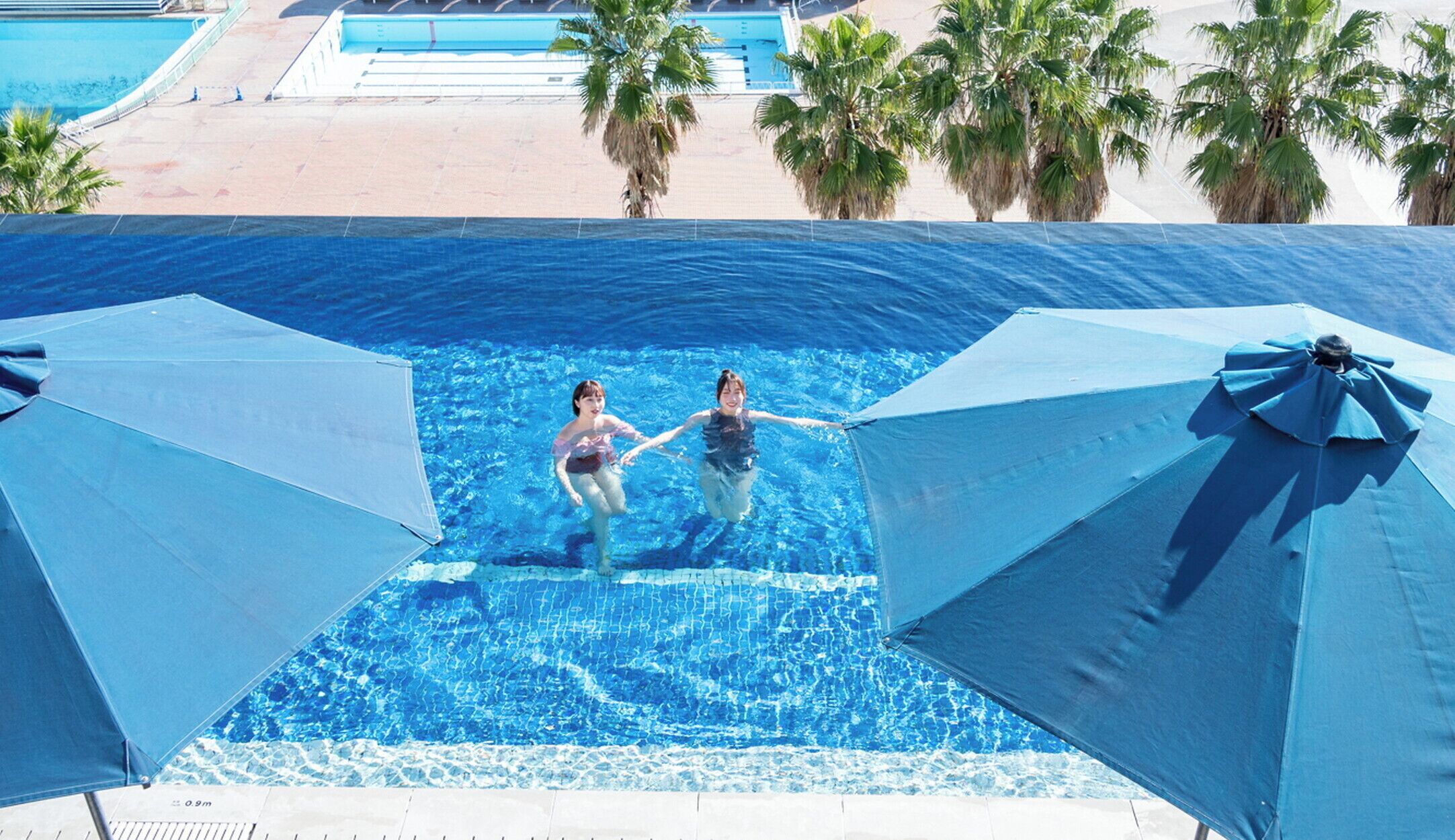 Une piscine où se trouvent deux personnes, entourée de palmiers et de parasols.
